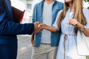 A young couple and agent examining a home's exterior and yard on a sunny day in a residential neighborhood.